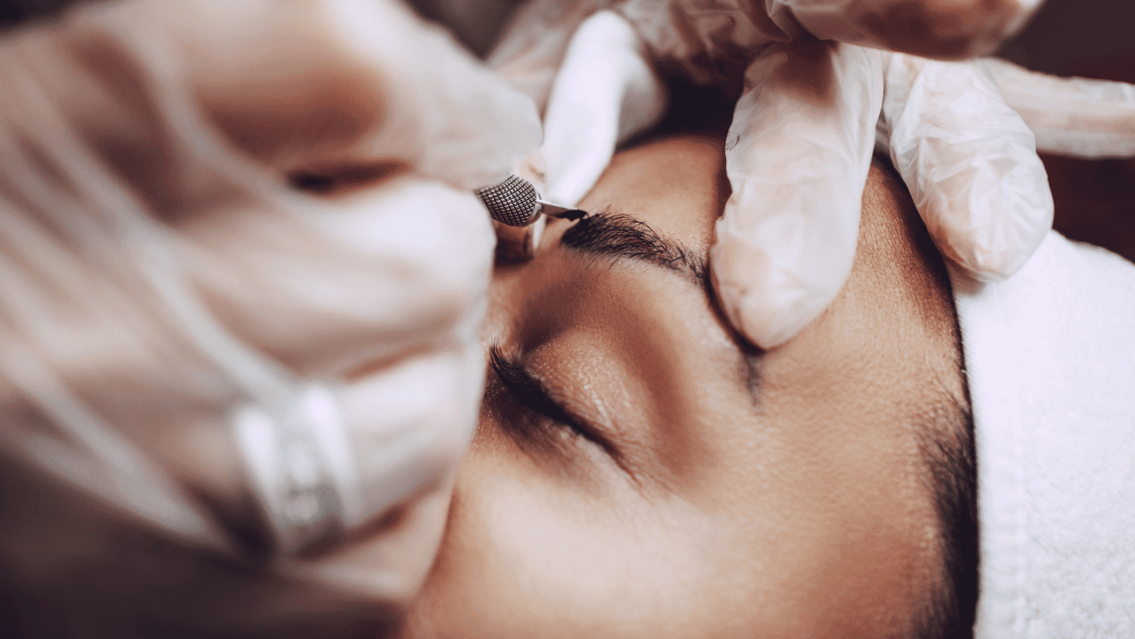 Person undergoing eyebrow microblading procedure with gloved hands and a specialized tool.